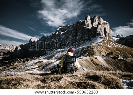 A person with a yellow jacket and black backpack sitting in front of a mountain rage. The scene is shot at the Gardena Pass in the Dolomite mountains, Northern Italy.