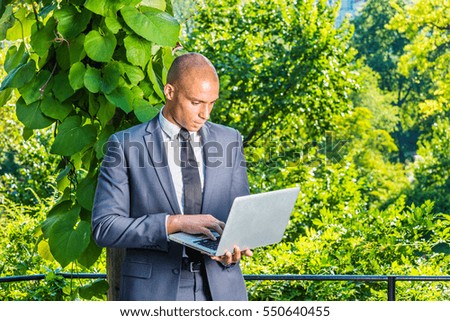 European Businessman traveling, working in New York. Wearing gray suit, black tie, Mixed Race French man with shaved head stands by big leaf plants, works on laptop computer. Color filtered effect.