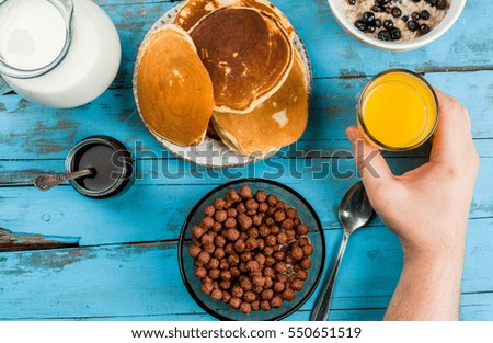 Man having breakfast with cereal chocolate balls, takes a glass of juice in hand. Pancake, jam and juice on table. View from above, hands in picture.