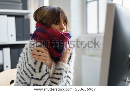 Sick businesswoman with winter chills and a fever sitting shivering in the office wrapped in a thick woolly winter scarf Royalty-Free Stock Photo #550656967