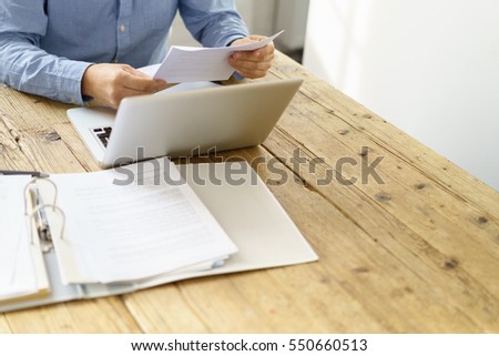 Businessman checking a typed report or document as he sits working on paperwork at a laptop computer, close up of his hands