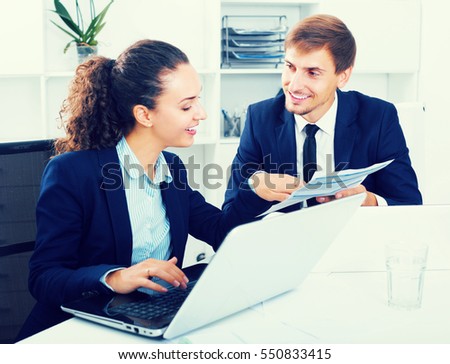 Portrait of positive adult business man and woman colleagues sitting with laptops on desk in office