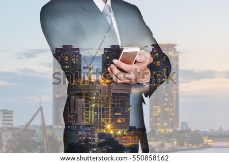 Double exposure of businessman hand hold smartphone, mobile, construction crane and building in the evening, twilight as business, technology, communication and industrial concept.