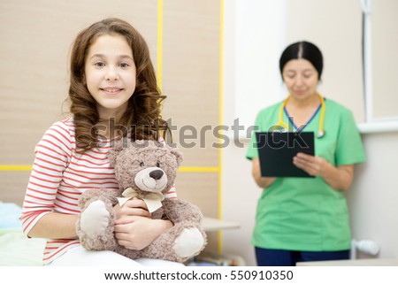 Happy healthy cutie. Beautiful young girl smiling to the camera holding her teddy bear sitting at the doctor's office her pediatrician writing notes on the background happiness vitality kids concept