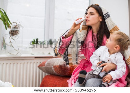 Young sick woman taking nose drops, sitting in sofa with little boy at home