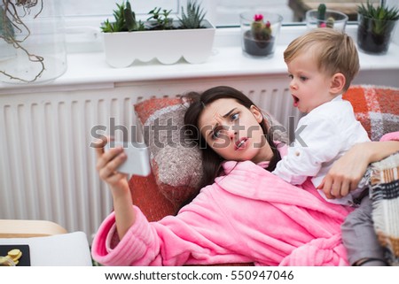 Sick young woman laying with little son in sofa, and making selfie on phone