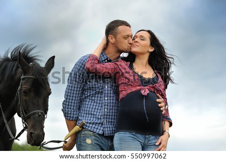 Awaiting the child. Young couple - she is a brunette with long hair, pregnant; he is tall and brave, holding the reins of the black horse, a walk on the ranch. He's kissing her