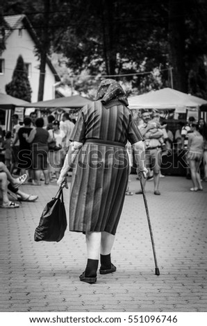 Old country lady walking and holding a cane and a bag in her hands with blurred people in background. Picture taken in black and white.
