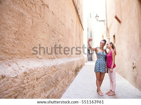 Two smiling teenagers girls friends visiting a destination city narrow street with stone buildings together, taking selfies pictures on student holiday, sunny outdoors. Travel technology lifestyle.