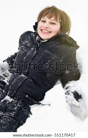 Happy young white girl lying in snow with smile.Portrait of funny chick in snowdrift.Warm winter clothes,black thick coat for cold weather.Happiness emotions on young woman face.White female model