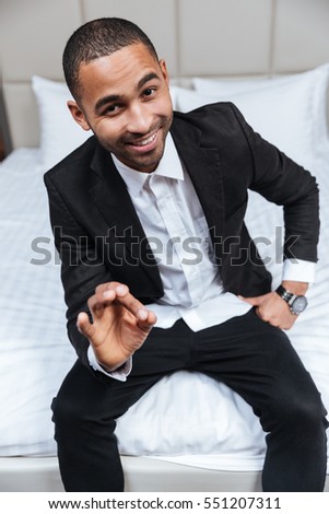 Vertical image of Smiling African man in suit sitting on bed in hotel room and showing ok sign