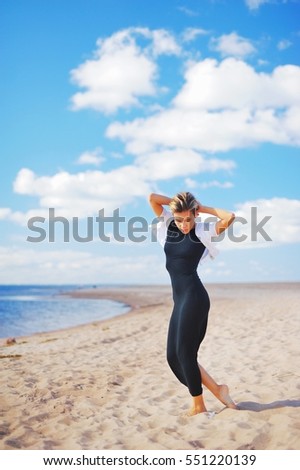 Pretty graceful girl in elegant black dress and white jacket is on the sandy beach, barefoot, his hands behind his head