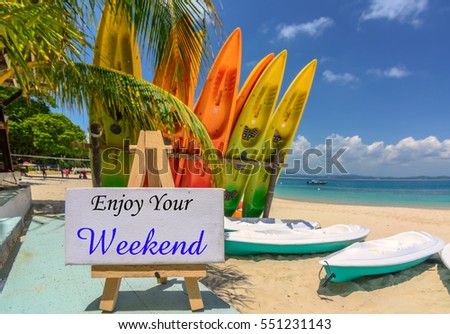 Conceptual image with word "ENJOY YOUR WEEKEND" on white canvas frame and wooden tripod. Blurred image of beach and colorful kayaks background. Image with selective focus