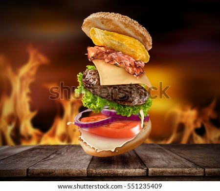 Maxi hamburger with flying ingredients placed on wooden planks with flames on background. Copy space for text, high resolution image