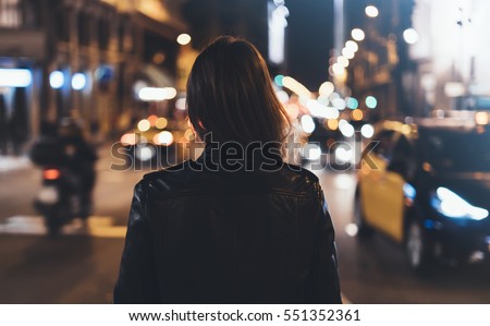 Hipster girl in black leather jacket from back on background illumination glow bokeh light in night atmospheric christmas city, hipster costs at a stop in headlights auto taxi; mockup glitter
