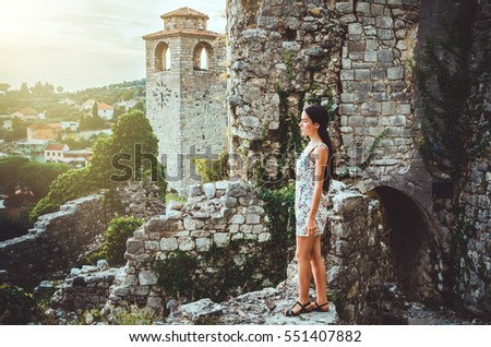 Feminine romantic woman posing in Stari Bar old fortress, Montenegro. Tanned female with long hair, red lips and manicure in white dress flowers. Brunette girl walks around oldest castle