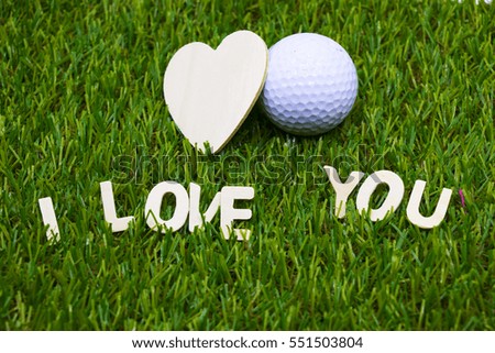Wooden heart shape and golf ball are on green grass.