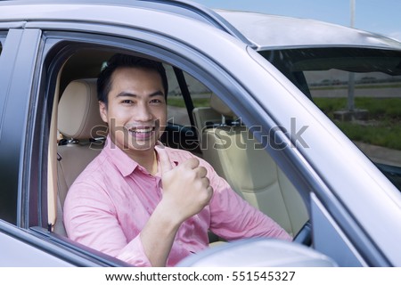 Image of happy young male driving a new car while showing thumb up and looking at the camera