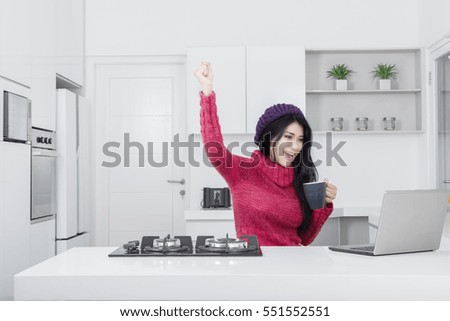 Picture of joyful young woman holding a mug while looking at the laptop and raise hands in the kitchen
