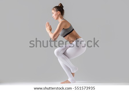 Young attractive woman practicing yoga, standing in Eagle exercise, Garudasana pose, working out wearing sportswear, white pants, gray top, indoor full length, isolated against grey studio background