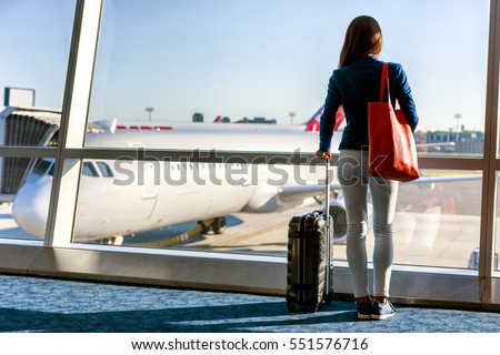 Travel tourist standing with luggage watching sunset at airport window. Unrecognizable woman looking at lounge looking at airplanes while waiting at boarding gate before departure. Travel lifestyle. Royalty-Free Stock Photo #551576716