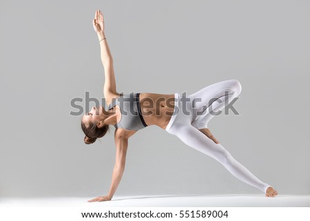 Young attractive woman practicing yoga, stretching in Side Plank exercise, Vasisthasana pose, working out wearing sportswear, white pants, indoor full length, isolated against grey studio background Royalty-Free Stock Photo #551589004