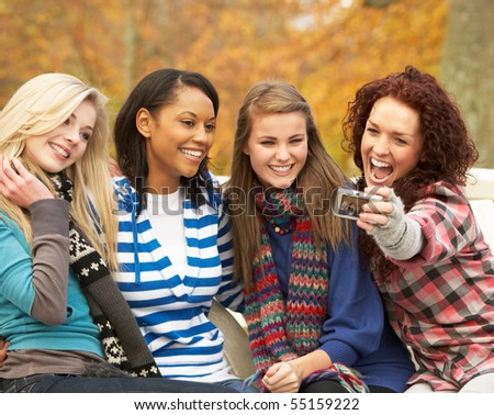 Group Of Four Teenage Girls Taking Picture With Camera Sitting On Bench In Autumn Park