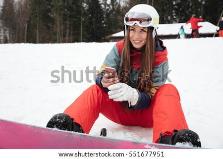 Photo of young happy lady snowboarder lies on the slopes frosty winter day while chatting by phone. Look at camera.