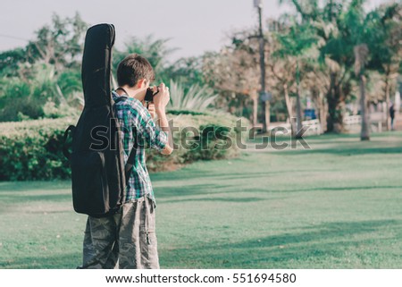 Boy slung guitar bag, taking picture in the garden with vintage camera.