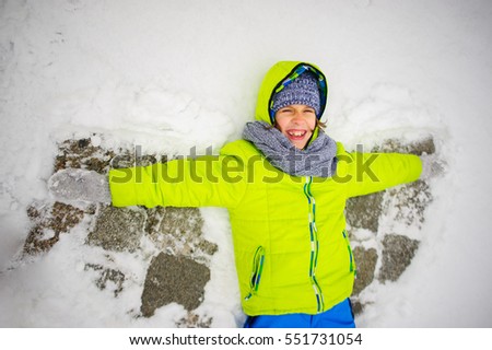 The cheerful boy of school age lies on snow and does wings of an angel. The boy is dressed in a bright ski suit. It is snowing. To the boy it is very cheerful.