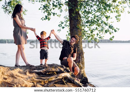 Young family with his young son is resting on the lake in the summer at sunset under a tree