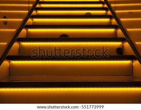 Yellow illuminated or neon-colored stairway, with yellow and orange colors.   Striking upward stairs with repeating horizontal lines. Urban background with mouseholes and led lighting.