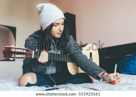Young beautiful eastern woman sitting on her bed in the bedroom holding guitar composing a song - musician, songwriter, composer concept  Royalty-Free Stock Photo #551923561