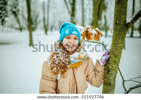 Little beautiful smiling girl in blue cap and beige jacket on background of snow-covered city park.