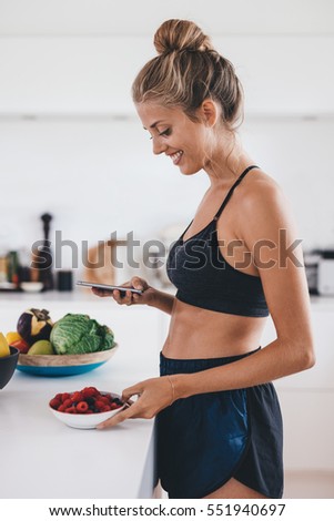 Side view of beautiful young woman standing in kitchen with bowl of fresh berries and reading text message on her mobile phone. Fit young woman in kitchen in morning.