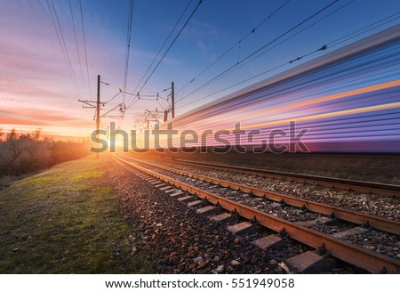 High speed passenger train in motion on railroad at sunset. Blurred commuter train. Railway station against sunny sky. Railroad travel, railway tourism. Rural industrial landscape. Concept Royalty-Free Stock Photo #551949058
