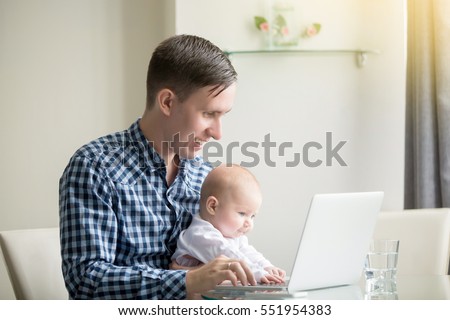 Young man working at desk at home with laptop, holding cute baby at knees, looking at screen, enjoying educational game, electronic babysitter, little helper, benefits of freelancing, safe computers
