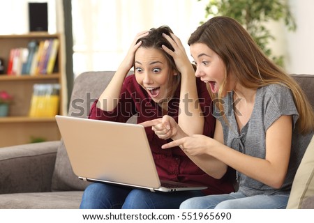 Two astonished roommates watching media content on line with a laptop sitting on a couch in the living room in a house interior Royalty-Free Stock Photo #551965690