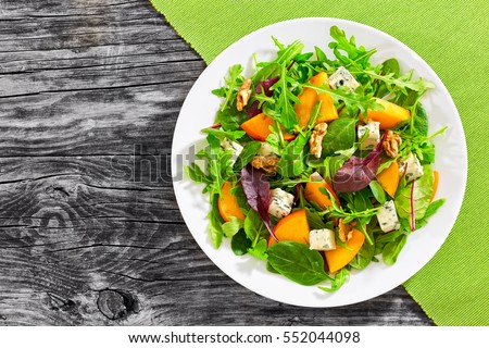 healthy delicious winter salad with persimmon slices, mix of spinach, arugula and lettuce leaves with blue cheese and walnuts on white plate on old dark wooden boards, view from above Royalty-Free Stock Photo #552044098