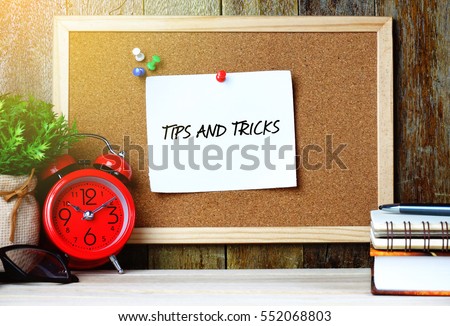 Paper note written with TIPS AND TRICKS inscription on cork board. Eye glasses, alarm clock, plant and books on wooden desk.