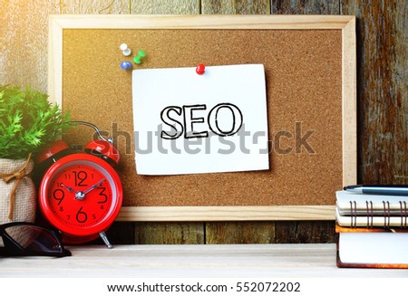 Paper note written with SEO text on cork board. Eye glasses, alarm clock, plant, pen and books on wooden table.