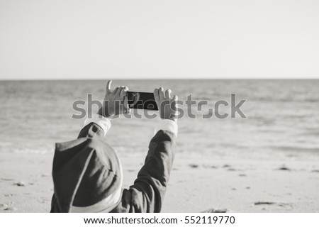 Child boy Santa Claus using mobile phone on ocean beach outdoors background, back view photo. Presents and gifts festive time new year holiday season. Black and white picture