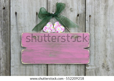 Weathered pink wood blank sign with flower hearts and green bow hanging on rustic antique wooden door; Valentine's Day home decor background with copy space