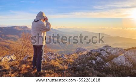 Traveller man in sports wear stands on the top of autumnal mountains with dry grass, sunset sky above and scenic landscape at the background taking photo by his camera.