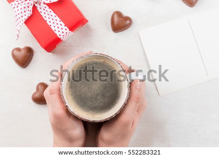 Concept for the celebration of Valentine's flavored coffee, candy heart shaped gift in red paper with ribbon and card. Space for text or congratulations. Hands in the frame, the girl holds a card 