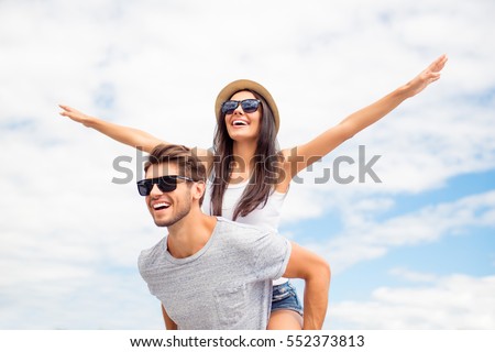 Young guy piggybacking cheerful girlfriend like airplane on the background of sky Royalty-Free Stock Photo #552373813