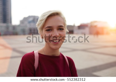Portrait of cheerful teenage female hipster student spending time outdoors walking on city street downtown with pink rucksack standing on blurred background copy space area for your advertising