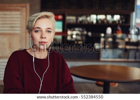 Portrait of attractive female hipster with natural makeup and trendy short haircut dressed in casual clothing sweatshirt listening to music via earphones sitting near  