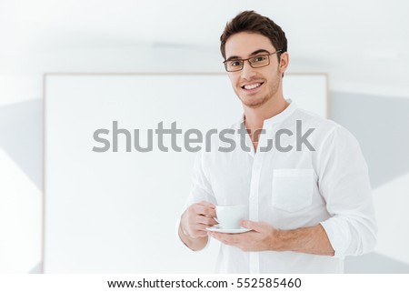 Photo of cheerful man wearing eyeglasses and dressed in white shirt holding cup of coffee near big board. Look at camera.