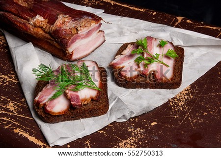 Close up view slices of smoked bacon with rye black bread on the white packaging paper. Composition in a rustic style on brown vintage background.